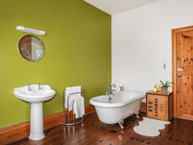 A bathroom with a white pedestal sink a round mirror a towel rack a clawfoot bathtub a wooden crate side table with a plant and candles and a wooden door at West Point Lynas Lighthouse Keeper's Cottage in Llaneilian near Amlwch
