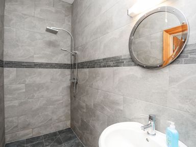 A bathroom with grey tiled walls a shower area a round mirror a sink and a soap dispenser at West Point Lynas Lighthouse Keeper's Cottage Llaneilian near Amlwch