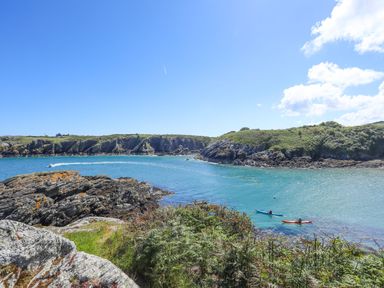 A coastal scene with rocky cliffs green vegetation two kayakers in the water and a motorboat in the distance at West Point Lynas Lighthouse Keeper's Cottage in Llaneilian near Amlwch