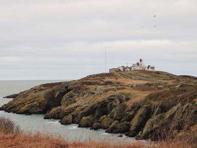 A coastal landscape with rocky cliffs and a cluster of buildings on a hill by the sea at West Point Lynas Lighthouse Keepers Cottage in Llaneilian near Amlwch