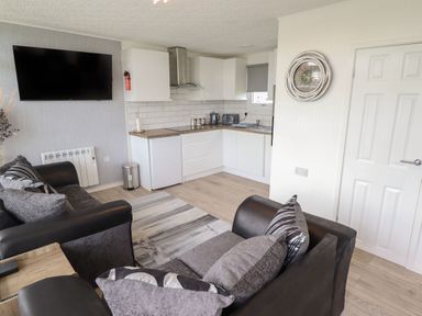 A living area with black and gray sofas a wall mounted tv a kitchen with white cabinets a small fridge and a circular clock on the wall at 39 Cherry Park in Chapel St Leonards