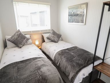 A bedroom with two single beds a window with blinds a small lamp an artwork on the wall and a shelving unit at 39 Cherry Park in Chapel St Leonards