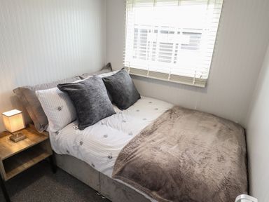 A small bedroom with a bed gray pillows bedside table with lamp and window blinds at 39 Cherry Park in Chapel St Leonards