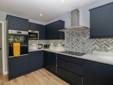A kitchen with dark cabinets a built in oven microwave stovetop and a backsplash with a herringbone pattern at Ranbir House in Longhope