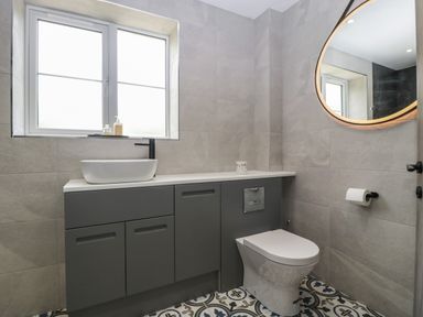 A bathroom with a gray cabinet, white sink, toilet, round mirror and window at Ranbir House in Longhope
