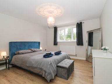 A bedroom with a blue bed frame gray bedding two bedside tables with lamps a window with black curtains a white dresser a tall mirror and a gray ottoman at Ranbir House Longhope