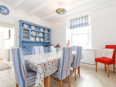 A dining room with a table covered by a patterned cloth surrounded by blue striped chairs and a blue cabinet with plates at Crab Pot Cottage in Porthleven