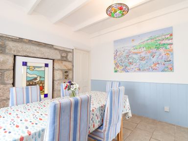A dining room with six striped chairs around a table with a patterned tablecloth and a stained glass artwork on a stone wall at Crab Pot Cottage in Porthleven