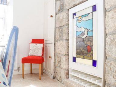A corner with a red chair and a nautical cushion next to a stained glass window set in a stone wall at Crab Pot Cottage in Porthleven