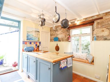 A kitchen with a blue island and hanging pans above a window seat with cushions at Crab Pot Cottage in Porthleven