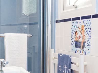 A bathroom with a glass shower door a white towel on a rack and decorative tiles with a painted scene at Crab Pot Cottage in Porthleven