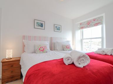 A bedroom with two single beds with red blankets and floral cushions next to a window with floral blinds at Crab Pot Cottage in Porthleven