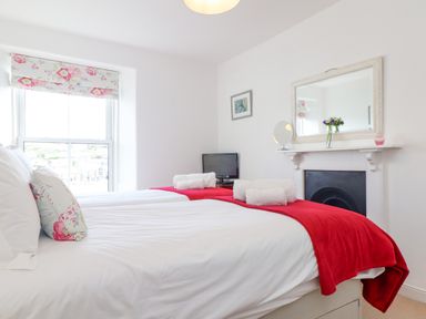 A bedroom with two beds side by side with white linens red throws and floral pillows a window with floral blinds a small television on a table and a mirror above a fireplace at Crab Pot Cottage in Porthleven
