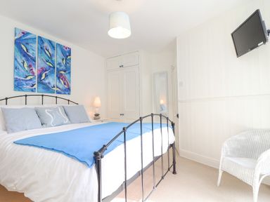 A bedroom with a metal bed frame blue bedding bedside table lamp wall art built in wardrobe armchair and wall mounted television at Crab Pot Cottage in Porthleven