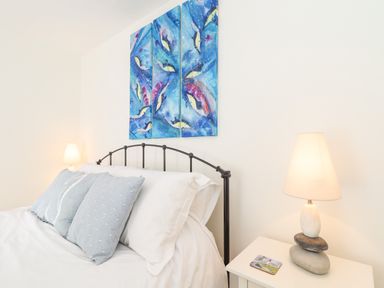 A bedroom with a bed with white sheets and blue pillows a bedside table with a lamp and a wall painting above the bed at Crab Pot Cottage in Porthleven