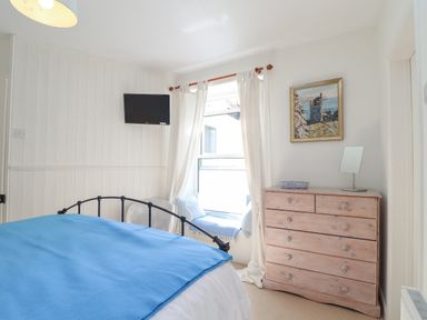 A bedroom with a blue blanket on the bed a wooden chest of drawers a television on the wall and a window seat at Crab Pot Cottage in Porthleven