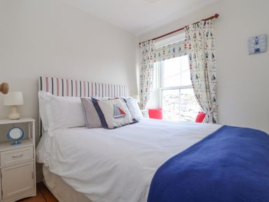 A bedroom with a bed with white bedding and striped headboard next to a window with patterned curtains at Crab Pot Cottage in Porthleven