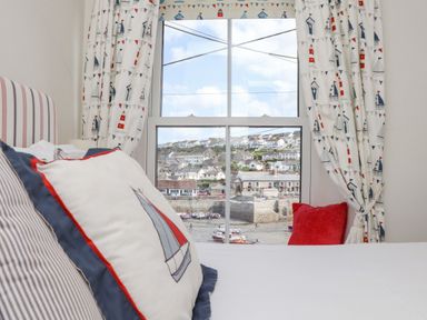 A bedroom window with patterned curtains and cushions overlooking a coastal village at Crab Pot Cottage in Porthleven