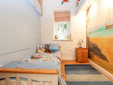 A small bedroom with a single bed with pirate-themed bedding a wooden nightstand with a lamp a window with a red striped blind and a mural of a beach scene on one wall at Crab Pot Cottage in Porthleven