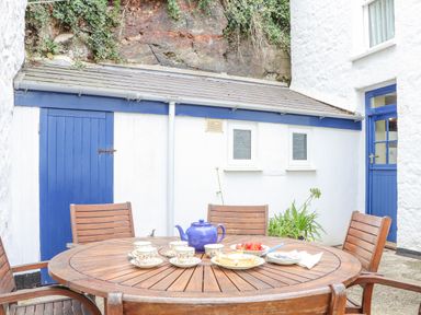An outdoor patio area with a wooden round table set with teacups plates and a teapot in front of white walls with a blue door at Crab Pot Cottage in Porthleven