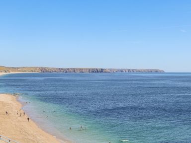 A sandy beach with people swimming and walking next to a large body of water and cliffs in the distance at Crab Pot Cottage in Porthleven