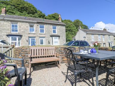 An outdoor patio with black metal chairs and table a wooden bench and stone houses behind at Crab Pot Cottage in Porthleven