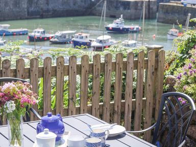 An outdoor table with tea set and flowers near a wooden fence overlooking boats on water at Crab Pot Cottage in Porthleven