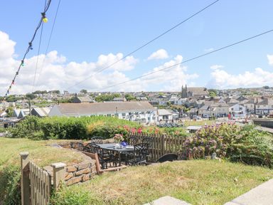 A garden patio with a table and chairs surrounded by greenery and flowers overlooking a village at Crab Pot Cottage in Porthleven