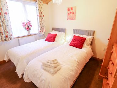 A bedroom with two single beds with white bedding and red pillows a wooden wardrobe and a window with floral curtains and flowers on the windowsill at Kantankye Lodge Thorpe St Peter near Wainfleet All Saints