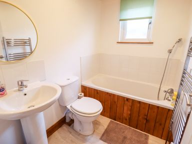 A bathroom with a pedestal sink, toilet, bathtub with wooden paneling, window with a green blind, and a wall-mounted towel radiator at Kantankye Lodge in Thorpe St Peter near Wainfleet All Saints