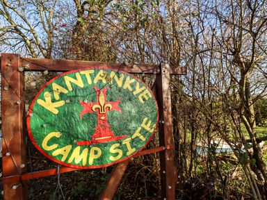 A wooden sign for Kantankye Camp Site with string lights in a wooded area at Kantankye Lodge Thorpe St Peter near Wainfleet All Saints