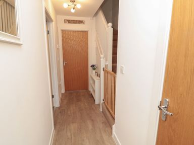 A hallway with a wooden floor a wooden door staircase with a safety gate and a small white shelf with a vase at Brucap Cottage in Beadnell