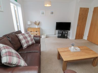A living room with a brown leather sofa with plaid cushions a wooden coffee table with a vase a TV on a stand and a cabinet with baskets at Brucap Cottage in Beadnell