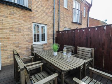 An outdoor patio with a wooden table and six wooden chairs a plant and a bottle with glasses on the table and a brick wall with windows at Brucap Cottage in Beadnell