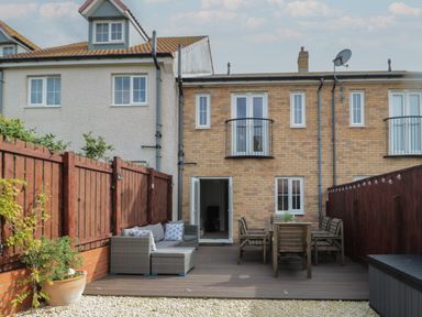 A backyard with wooden deck furniture and a brick house with a balcony at Brucap Cottage in Beadnell