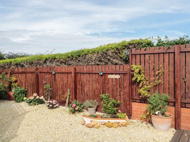A gravel garden area with potted plants and flowers beside a wooden fence with a sign pointing to coast path Beadnell at Brucap Cottage in Beadnell