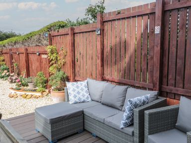 An outdoor seating area with gray cushioned wicker furniture and patterned pillows beside a wooden fence with plants and a gravel garden at Brucap Cottage in Beadnell
