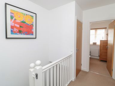 A hallway with white walls a staircase with white banisters a colorful framed painting and two wooden doors one open showing a room with a wooden chest of drawers at Brucap Cottage in Beadnell