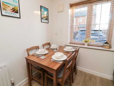 A dining area with a wooden table set for four near a window with blinds and two framed pictures on the wall at Brucap Cottage in Beadnell