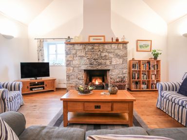 A living room with a stone fireplace a wooden coffee table striped armchairs a television bookshelf and wooden flooring at Rhyd Y Bont Bach in Rhoscolyn