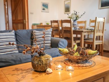 A living room with a blue sofa and striped pillows wooden dining table with chairs and a centerpiece with pine cones and pears at Rhyd Y Bont Bach in Rhoscolyn