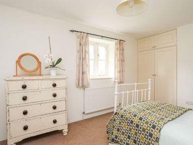 A bedroom with a white bed frame a patterned blanket a wooden dresser with a mirror a potted plant curtains and built in wardrobes at Rhyd Y Bont Bach in Rhoscolyn