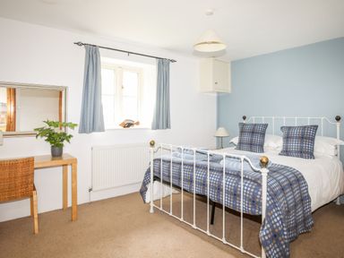 A bedroom with a white metal bed, blue patterned bedding, a wooden desk with a plant, a window with blue curtains at Rhyd Y Bont Bach in Rhoscolyn