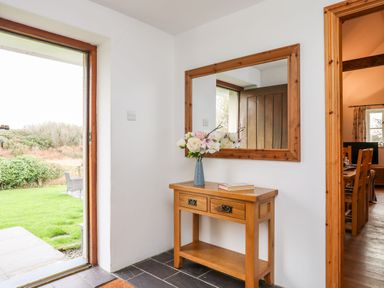 An entryway with a wooden table holding a vase of flowers and a book below a wooden framed mirror near an open door showing a garden at Rhyd Y Bont Bach in Rhoscolyn