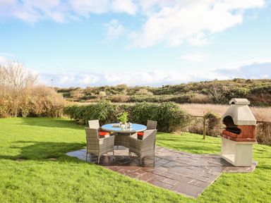 An outdoor patio area with a round table and four chairs next to a barbecue on a grassy lawn at Rhyd Y Bont Bach in Rhoscolyn