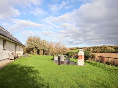 A backyard with green grass a patio area with table and chairs and a barbecue at Rhyd Y Bont Bach in Rhoscolyn