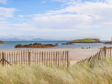 A beach scene with sand dunes, wooden fence, rocky shoreline, people near the water, and an island in the distance at Rhyd Y Bont Bach in Rhoscolyn