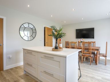 A kitchen island with a vase and bottle on it a wooden dining table with chairs and a wall-mounted tv in Iscoed Llangefni