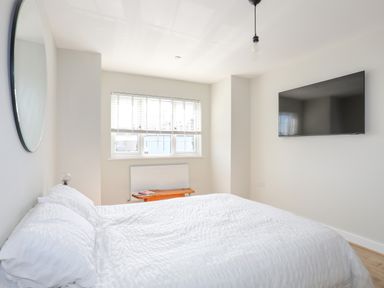 A bedroom with a bed white bedding a round mirror a window a bench and a wall mounted television at Iscoed in Llangefni