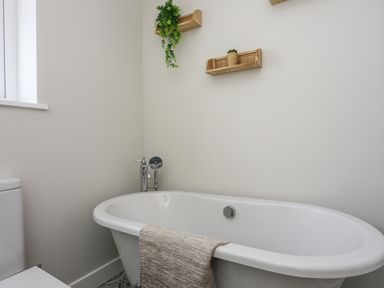 A bathroom with a white bathtub with silver fixtures a towel hanging over the tub and small wooden shelves with plants on the wall at Iscoed in Llangefni